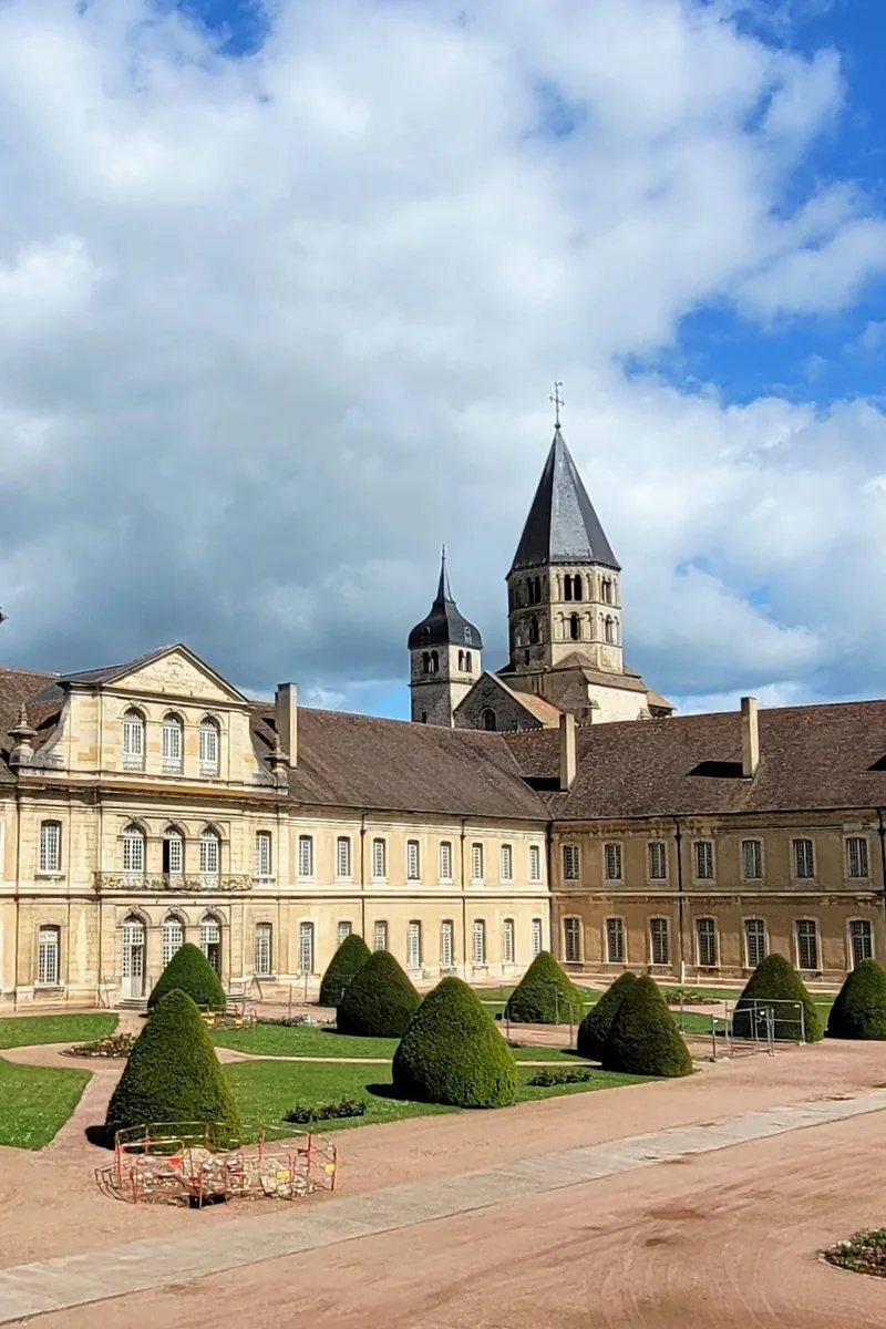 The 18th-century abbatial palace of the Abbey of Cluny with its formal topiary courtyard, and the octagonal Clocher de l'Eau Bénite — the surviving Romanesque bell tower of the great abbey church — rising behind under a blue Burgundy sky. Cluny, France.